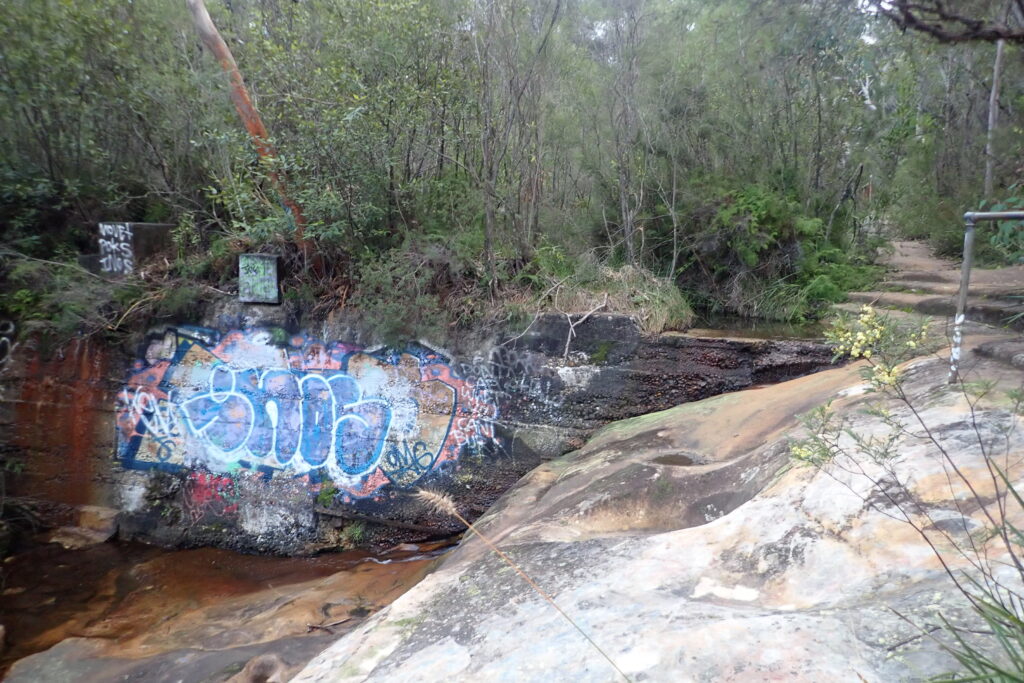 Looking up creek towards graffitied retaining wall, steps and track.